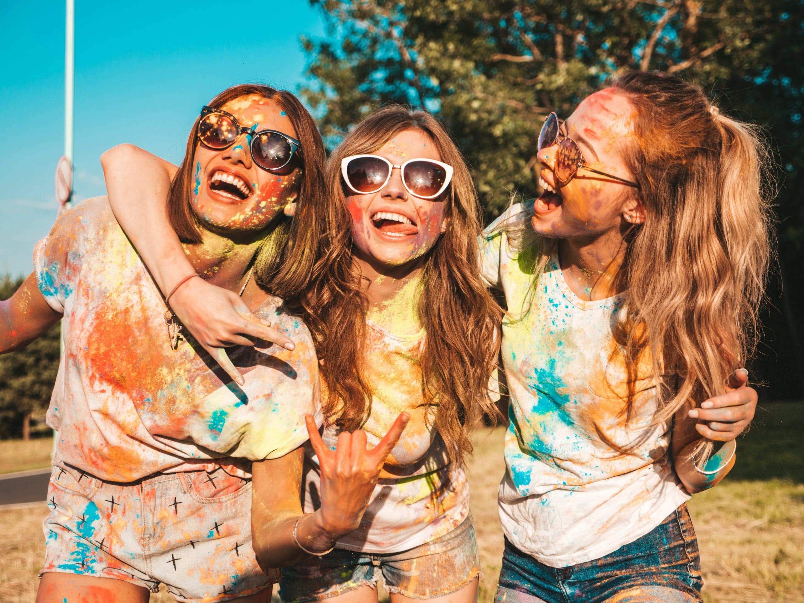 Three happy beautiful girls making party at Holi colors festival in summer time.Young smiling women friends having fun after music event at sunset. Positive models going crazy in sunglasses at sunset